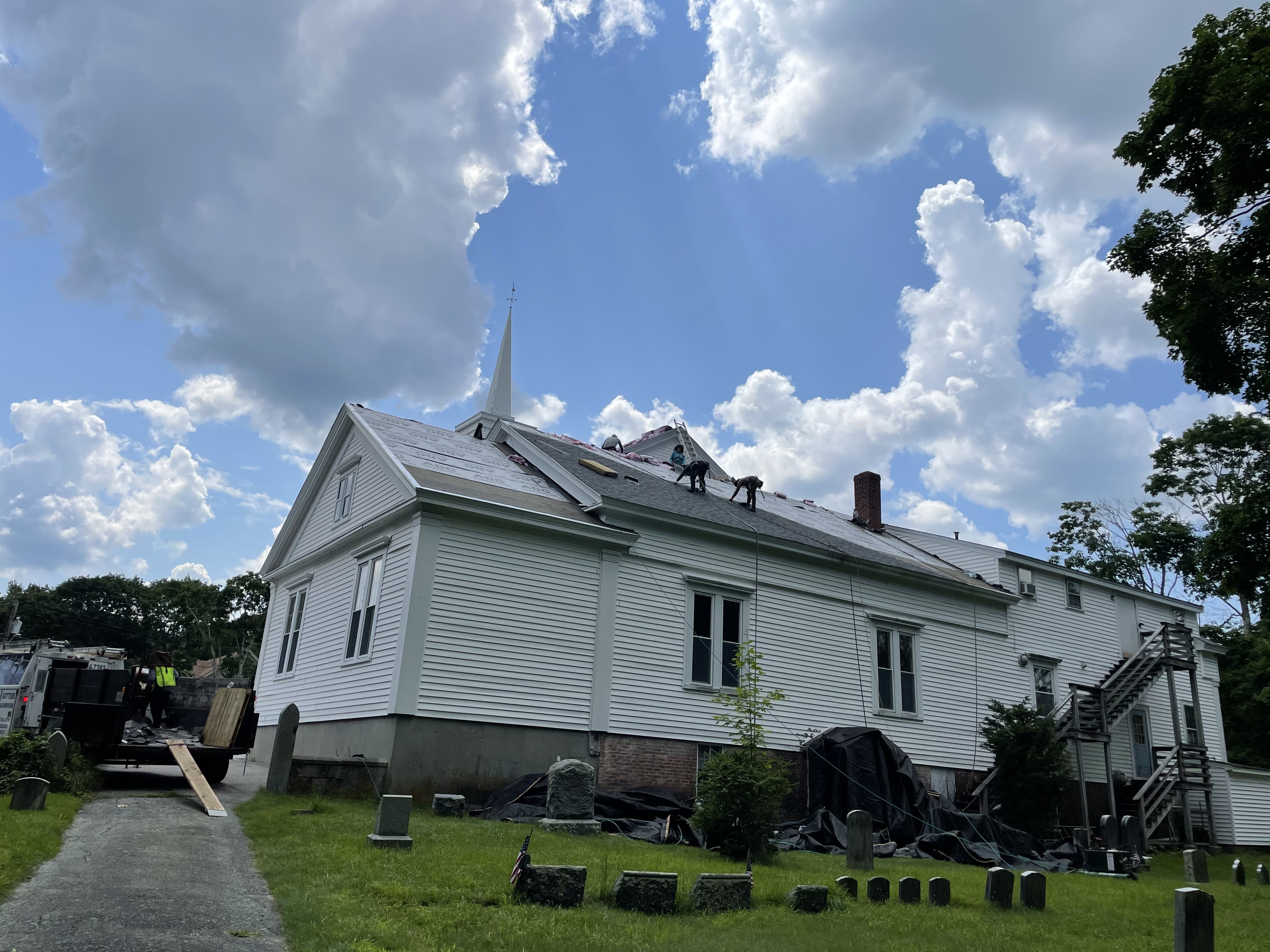 White steepled church in East Bridgewater, MA with Storm King Roofing crew completing the roof replacement and a dump trailer on site