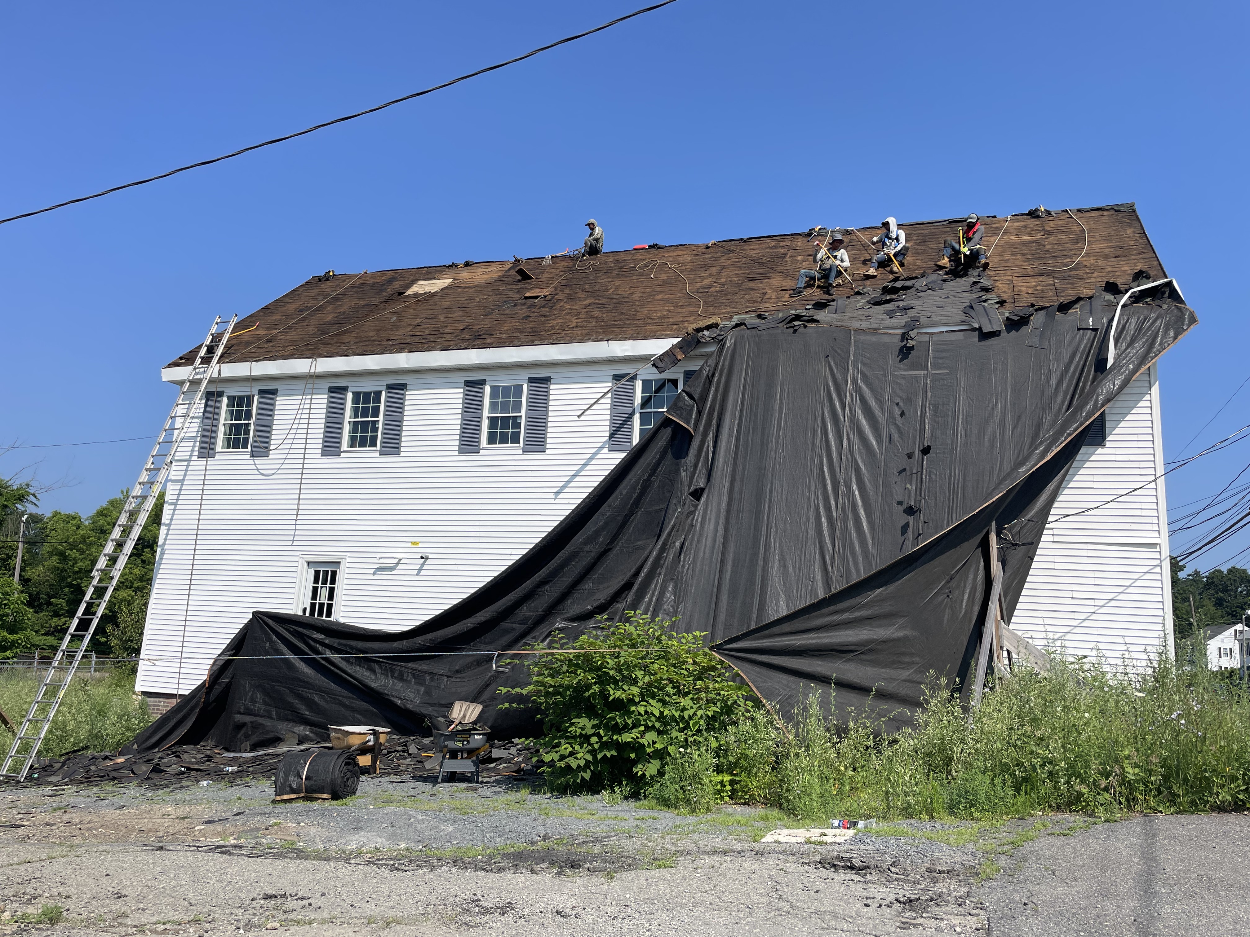 Storm King Roofing crew on the stripped wood deck of a large white building in East Bridgewater, MA with old shingles tarped down the gable end during a full tear-off