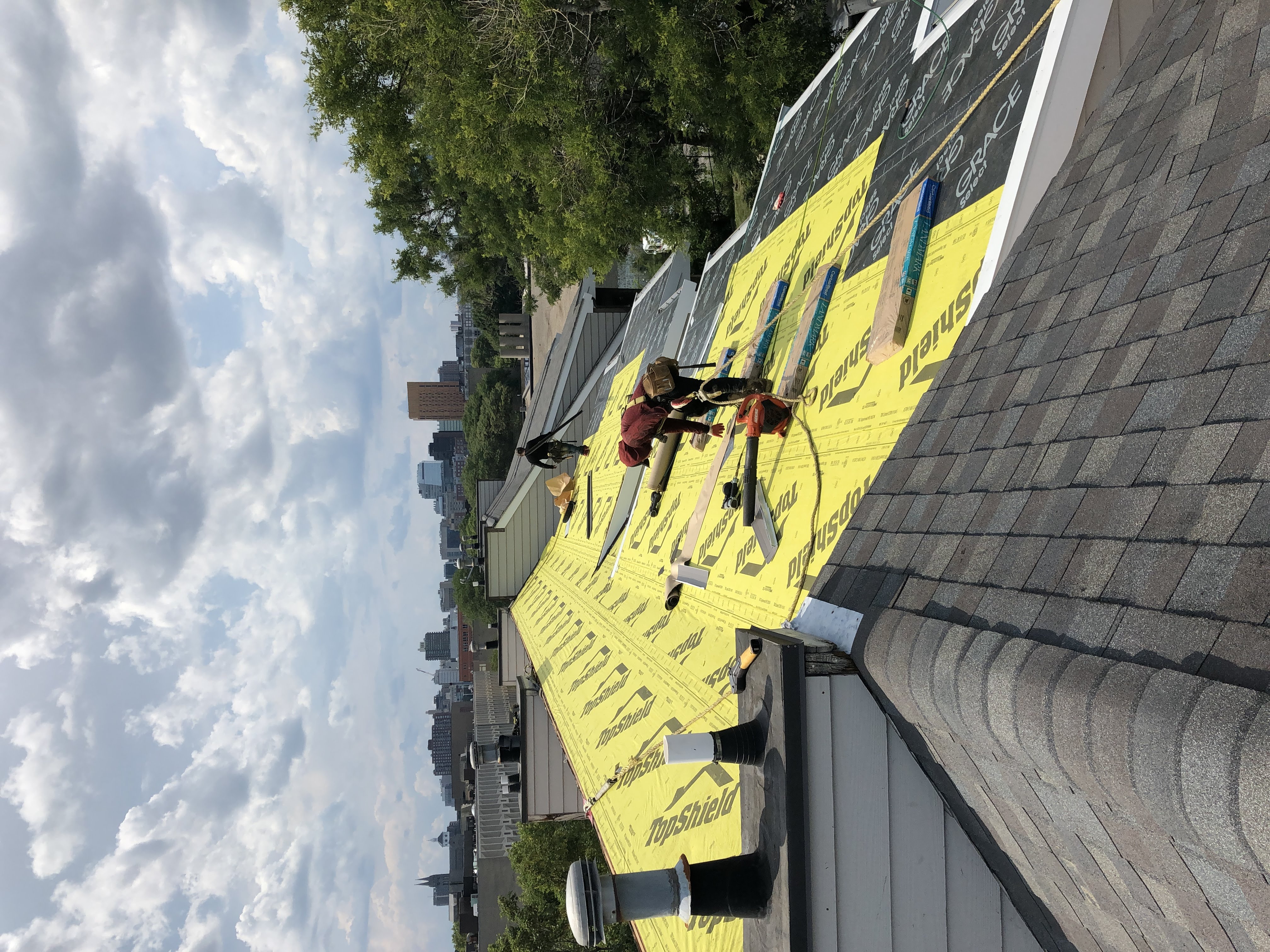 Storm King Roofing worker installing shingles over yellow TopShield underlayment on a Brookline, MA home with the Boston skyline in the background