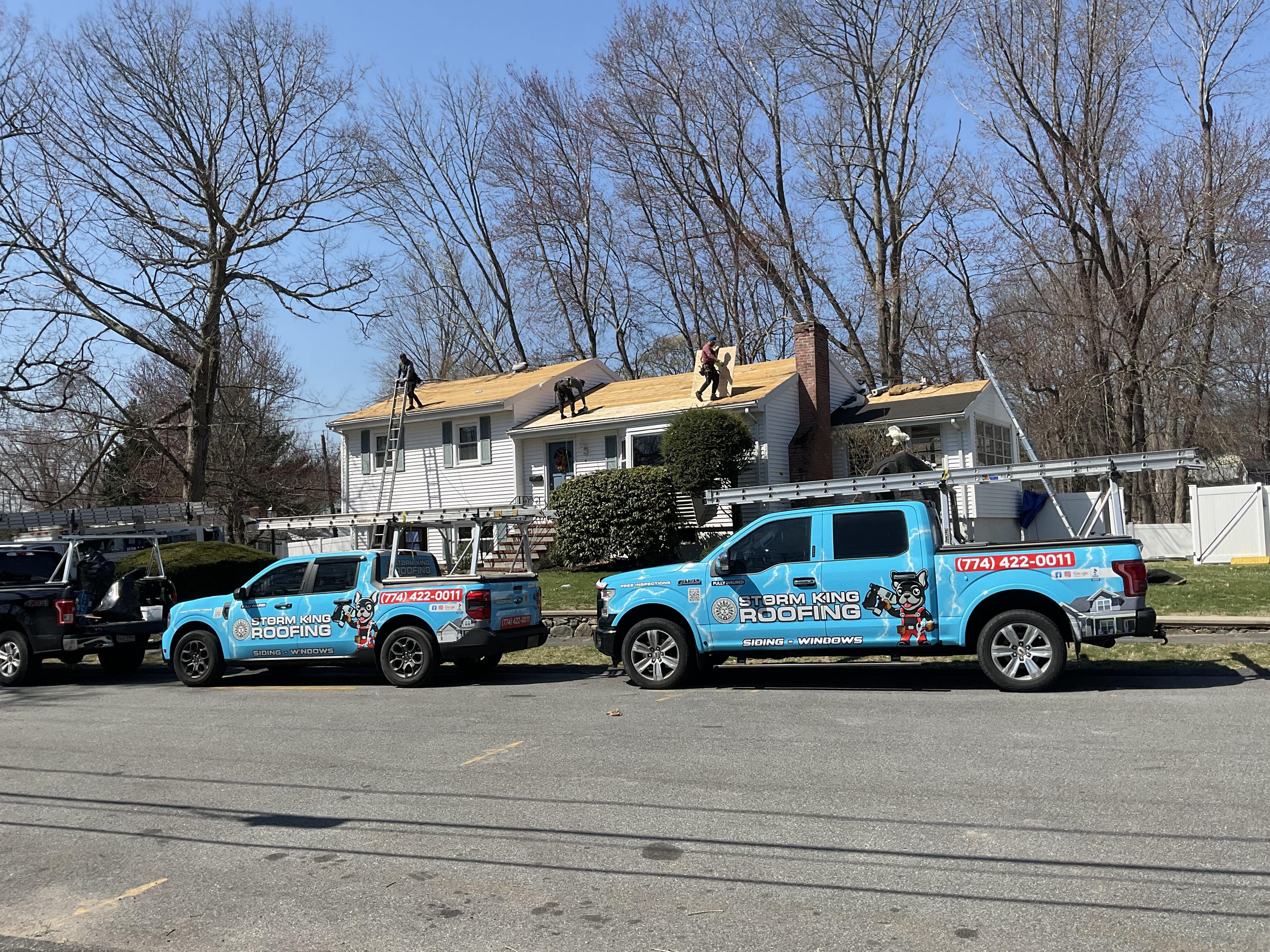 Two branded Storm King Roofing trucks parked while the crew tears off and replaces a shingle roof in Norwood, MA